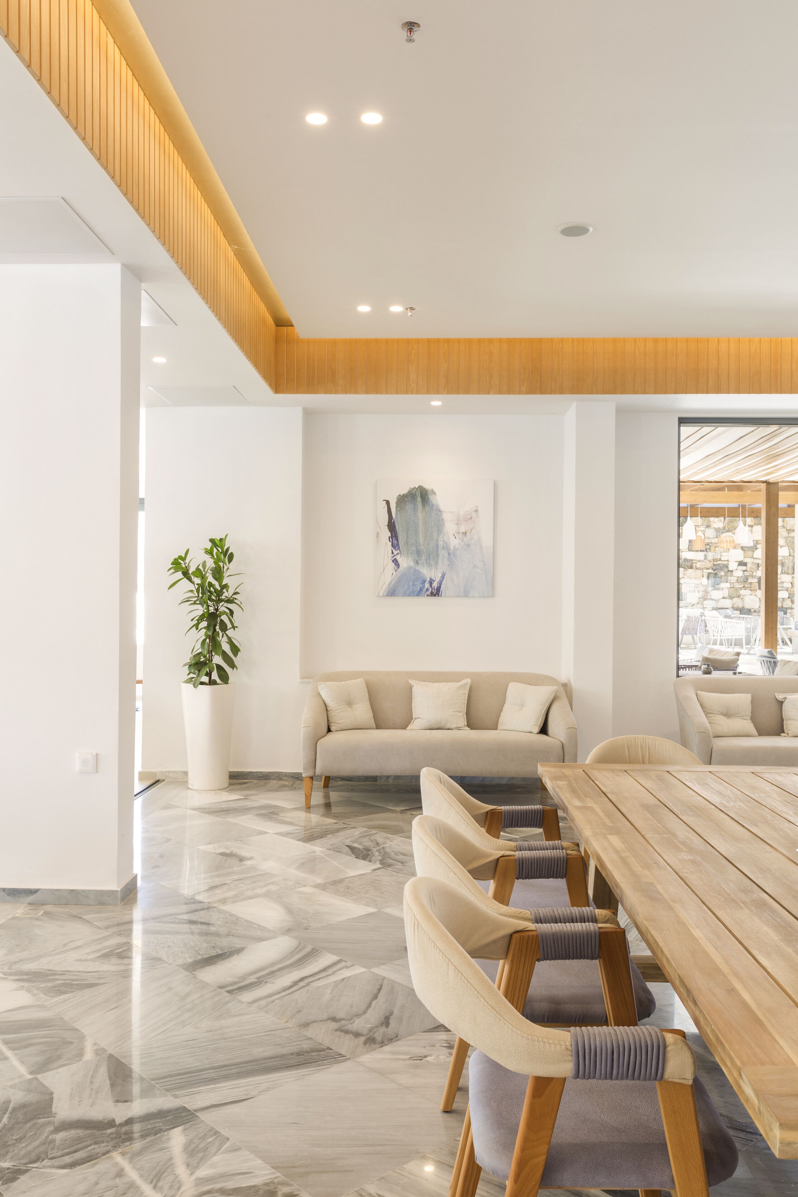 Un comedor moderno con una mesa de madera y sillas acolchadas, una planta en maceta y un sofá color beige.