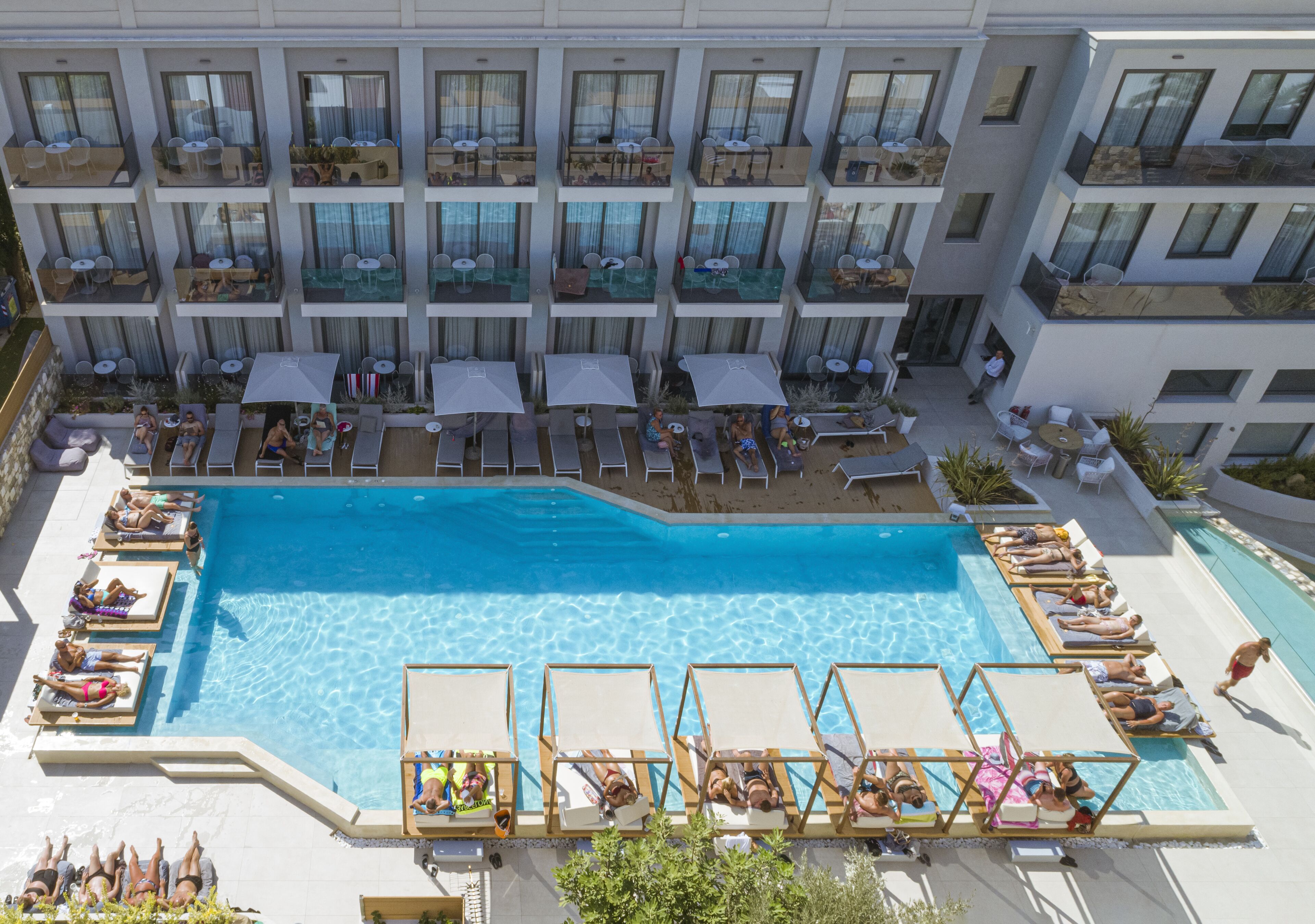 Ein Poolbereich auf dem Dach mit Liegestühlen und Sonnenschirmen, umgeben von einem mehrstöckigen Gebäude mit Balkonen.