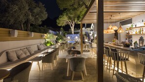 An outdoor dining area with wicker furniture, a bar with shelves of bottles, and a view of trees and mountains in the background. - Samian Mare Hotel Suites & Spa (Samos)
