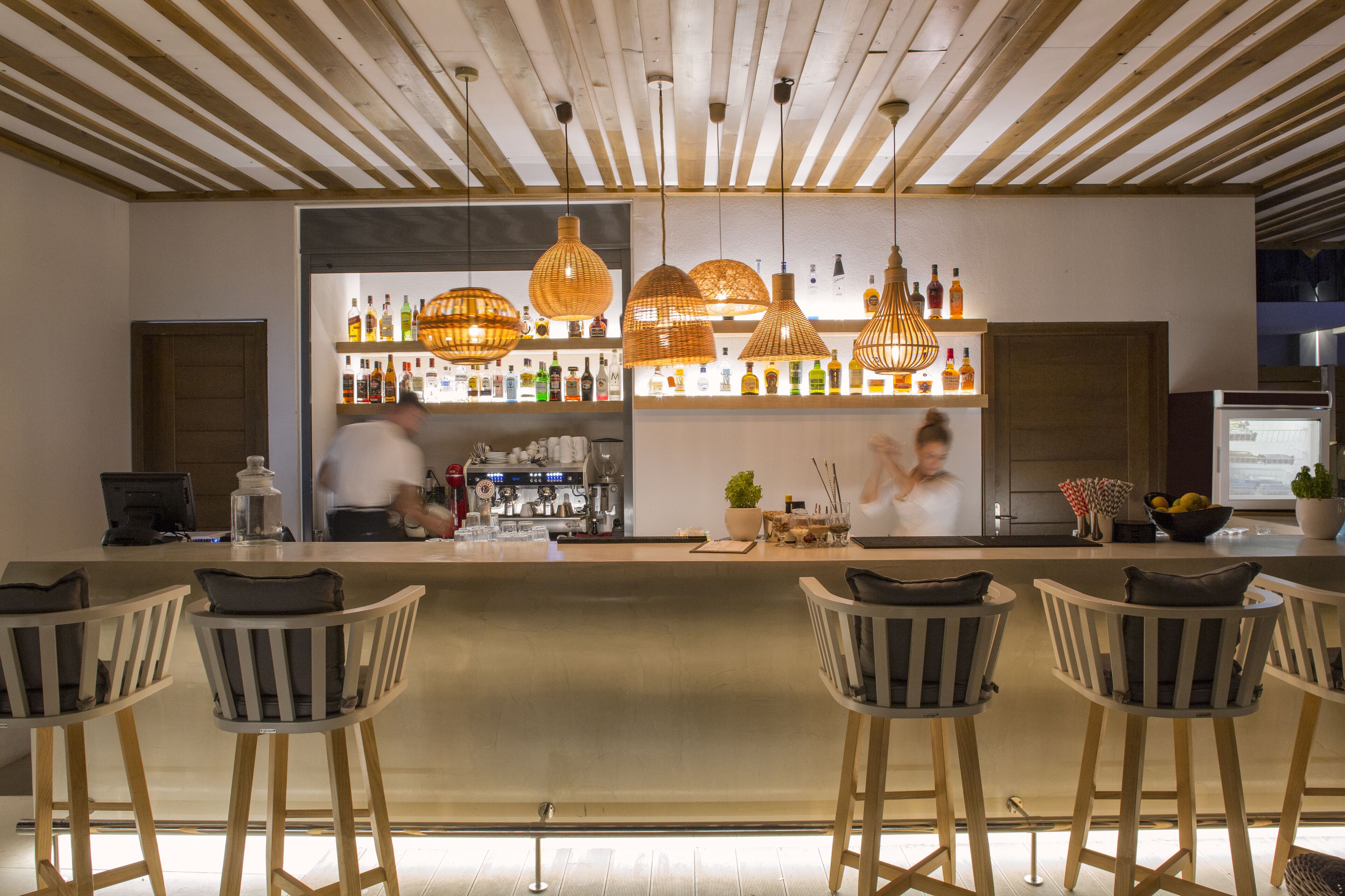 A bar area with wooden ceiling beams, hanging wicker light fixtures, and a bar counter with a variety of bottles. There are four bar stools with cushions.