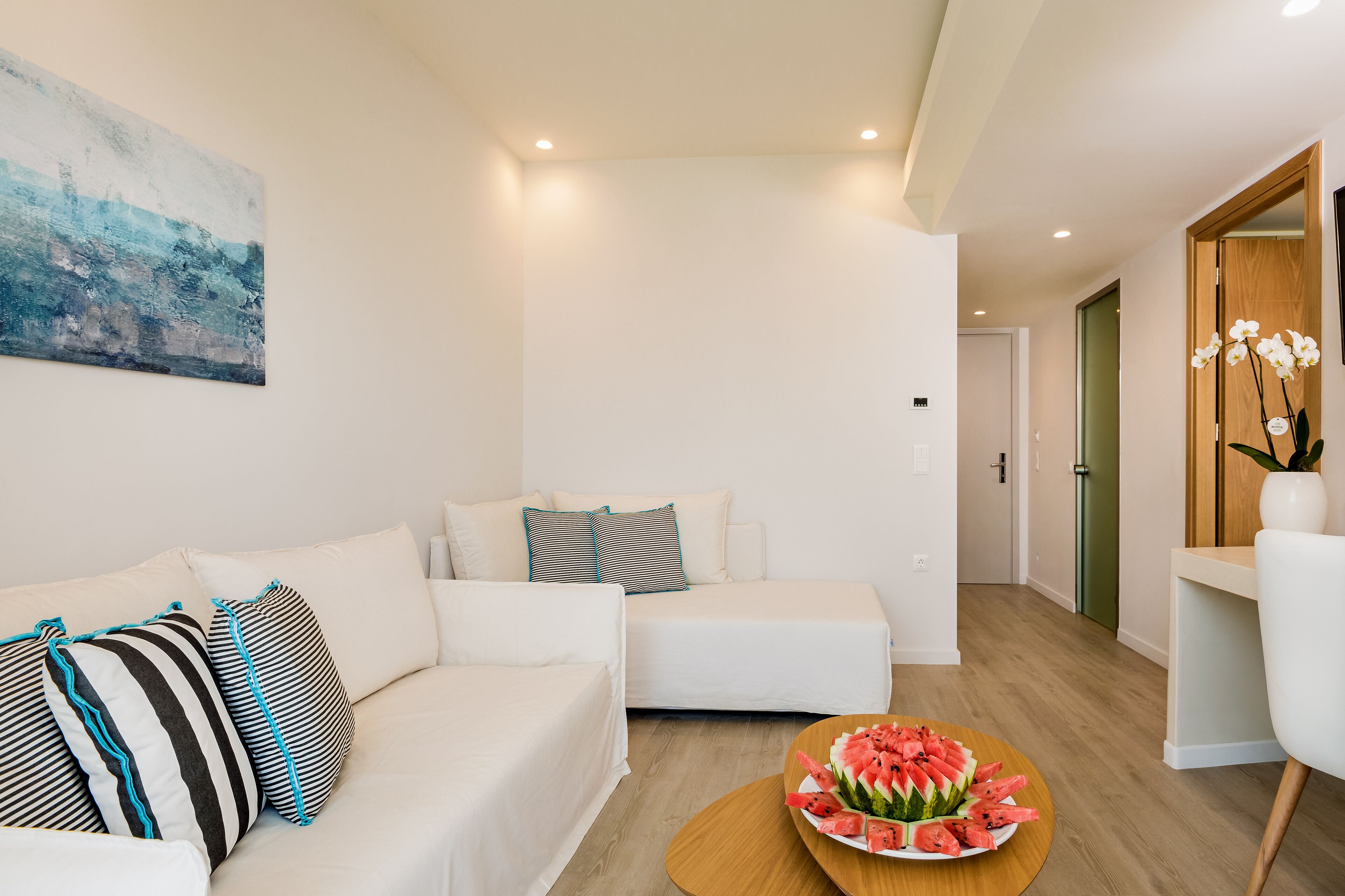 Una sala de estar moderna con un sofá blanco, cojines a rayas, una mesa de centro de madera con una sandía como adorno central y un cuadro en la pared.