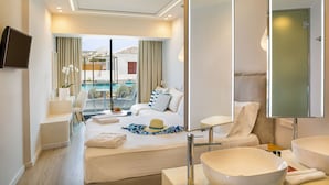 A hotel room with a bed, a large mirror, a TV, a bathroom with a large sink, and a view of a pool and building outside. - Samian Mare Hotel Suites & Spa (Samos)