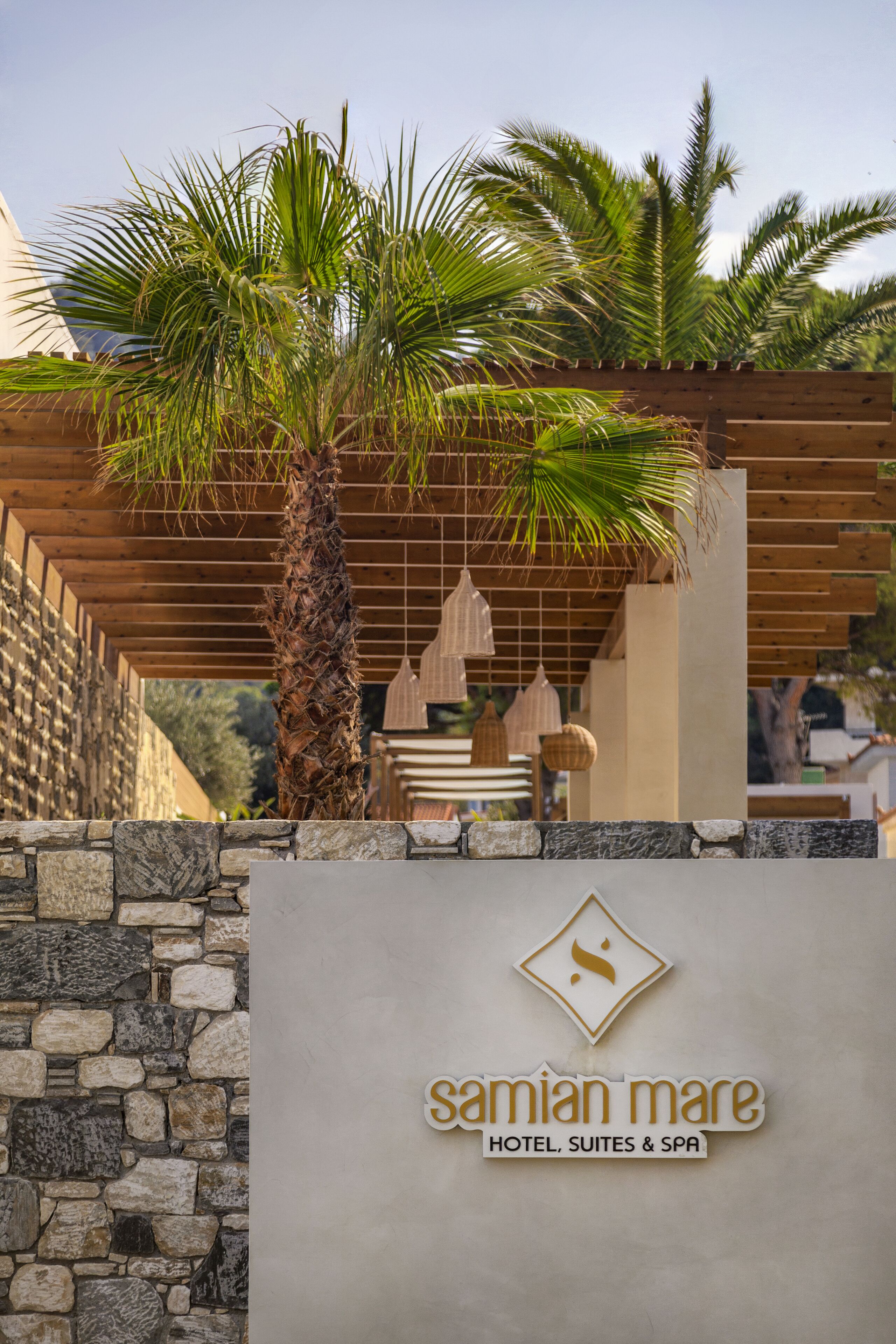 A stone wall with a wooden pergola and palm trees. The wall has a sign that reads "Samian Mare Hotel, Suites & Spa".