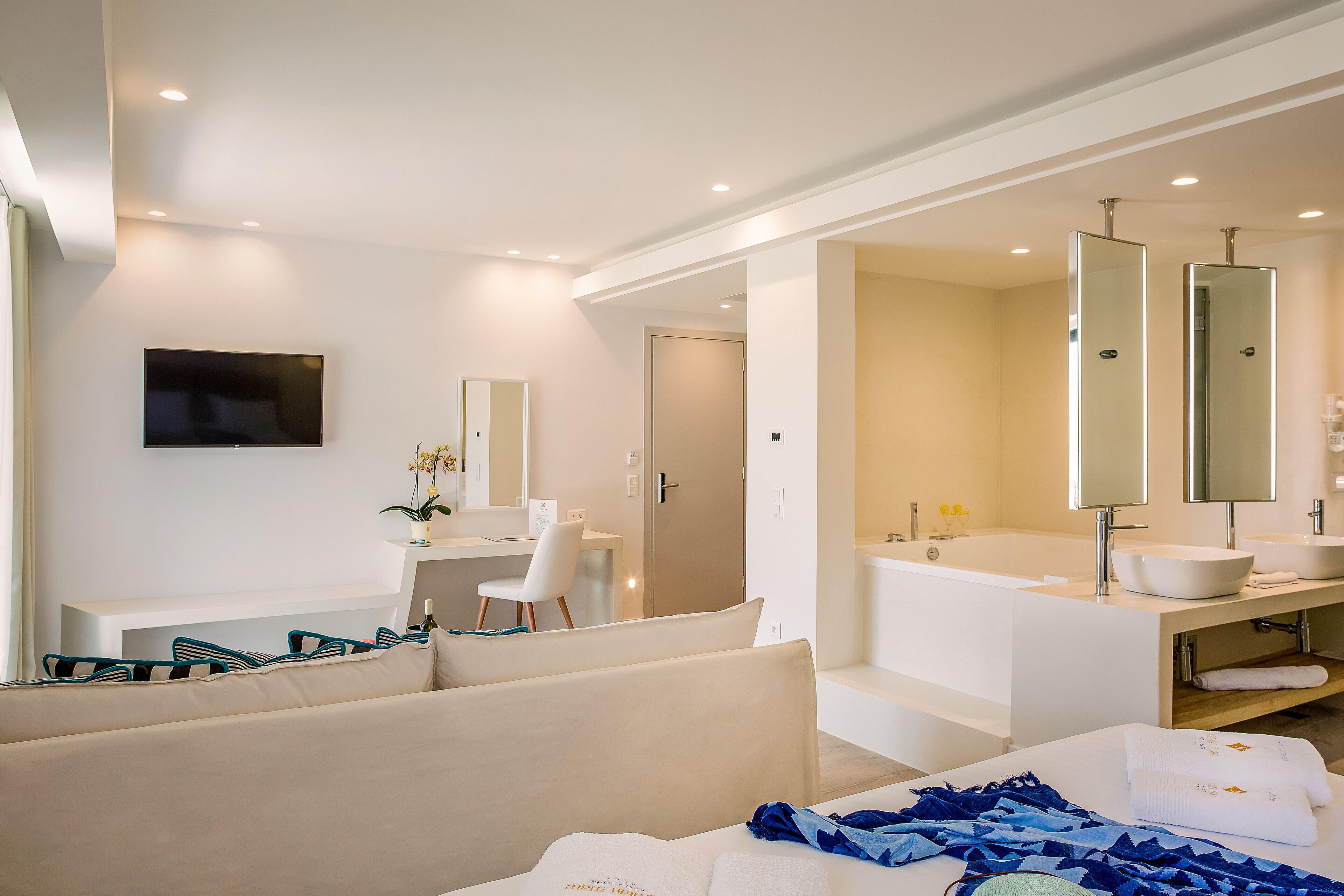 A modern hotel room with a sofa, a desk, a flat-screen TV, a large mirror, a bathtub, and a vanity with a sink.