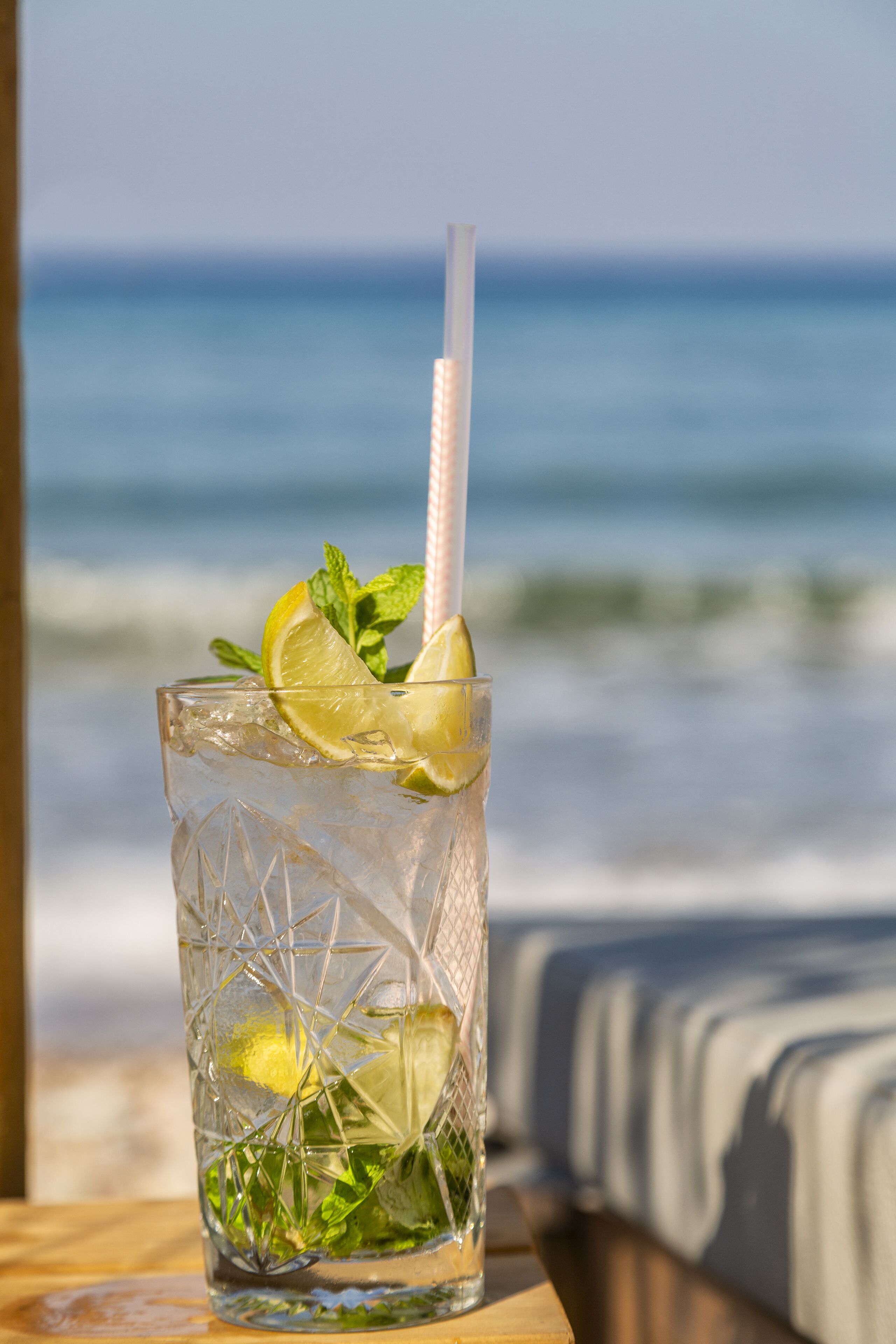 A clear glass with a straw, lime slice, and mint garnish on a wooden table with a beach view in the background.