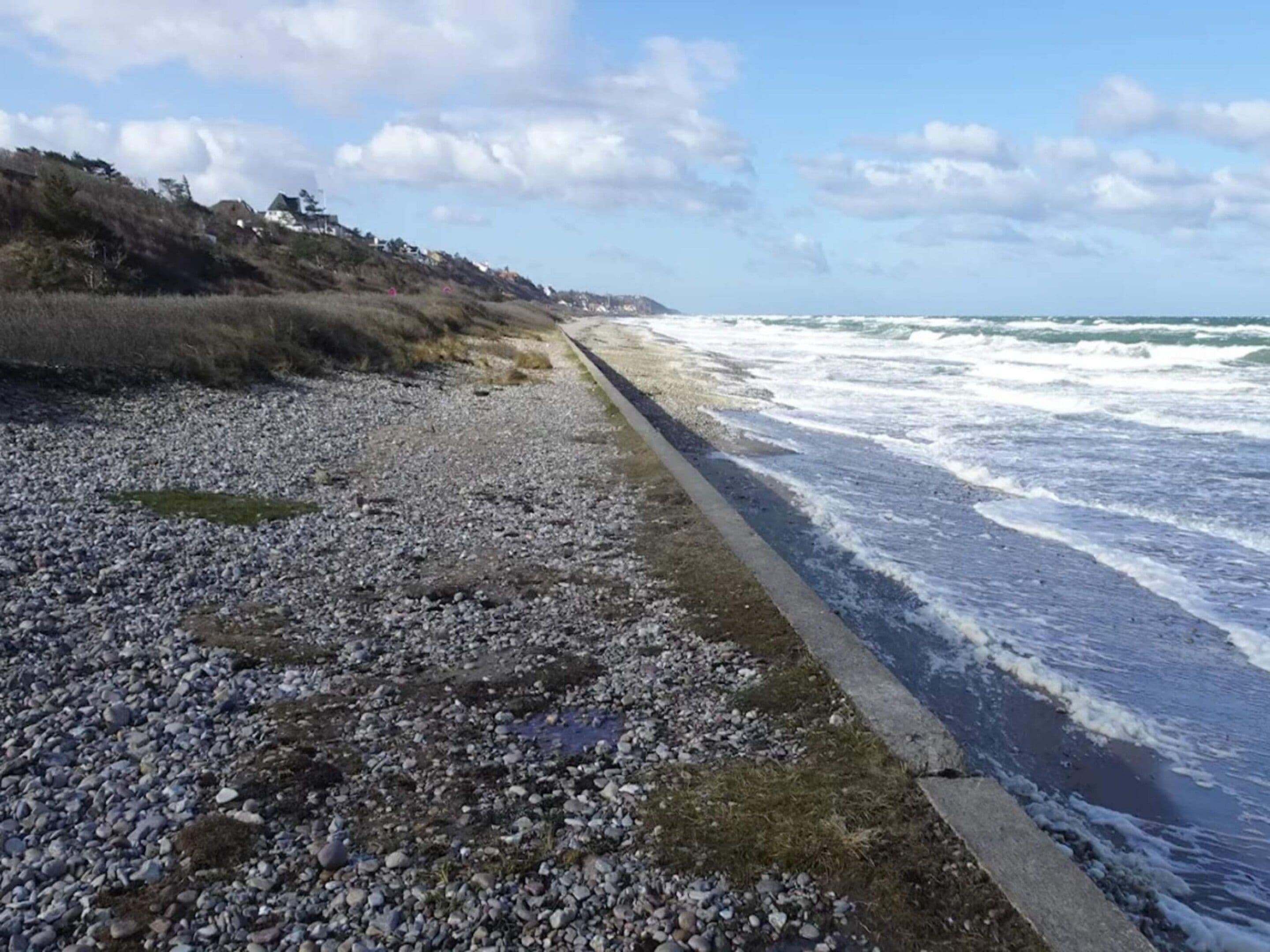 A rocky beach with waves crashing onto the shore, a concrete path along the water, and a hilly landscape in the background.