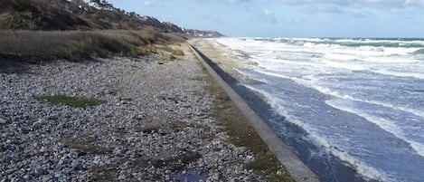 A rocky beach with waves crashing onto the shore, a concrete path along the water, and a hilly landscape in the background.