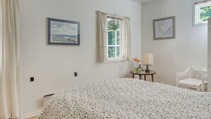 A bedroom with a bed, two windows, a chair, and a small table with a lamp.