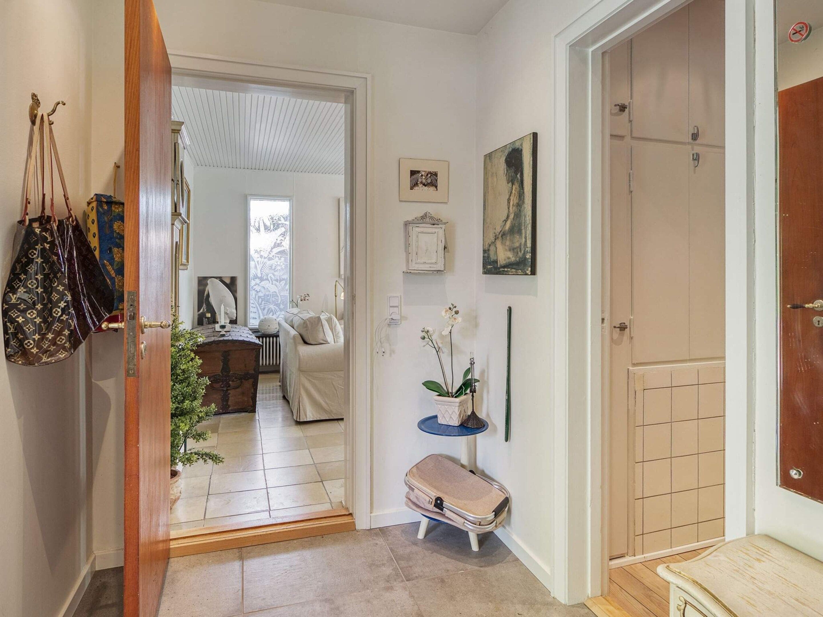 Un pasillo con una puerta que da a una habitación, una mesa pequeña con un florero y una maleta en el suelo.