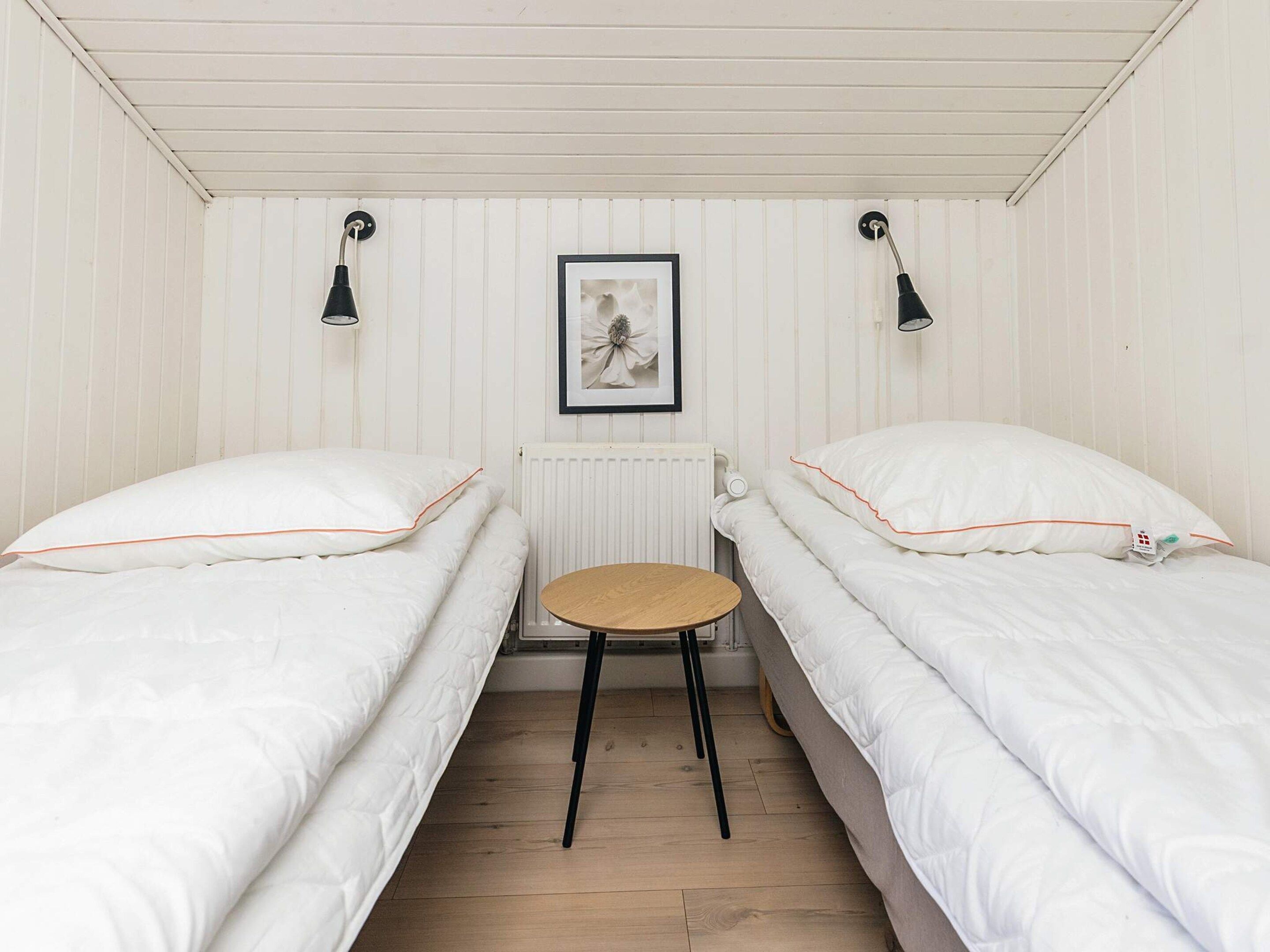 A small, single-room accommodation with two beds, a round table, and a framed picture on the wall.