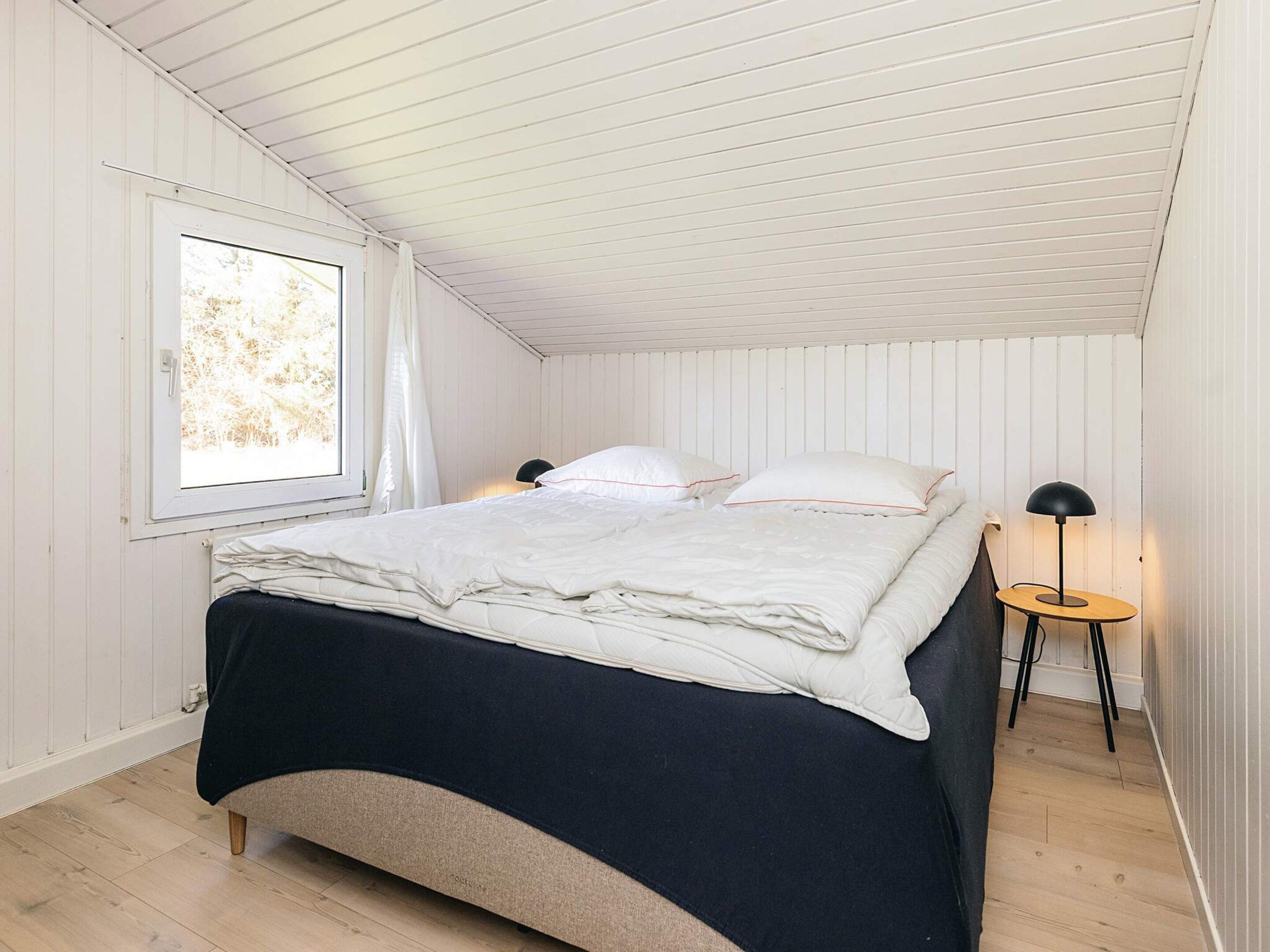 A bedroom with a bed, a window, a bedside table with a lamp, and a wooden floor.