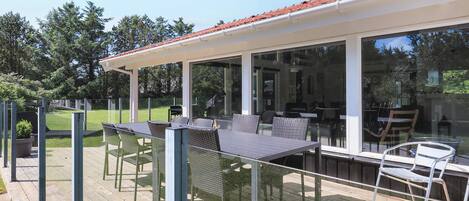 An outdoor deck with a dining area, including tables and chairs, and a glass enclosure leading to a building.