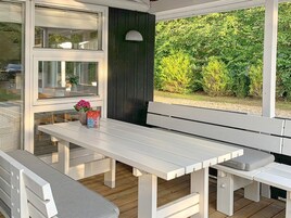 Zona de estar al aire libre techada con bancos blancos y una mesa, rodeada por puertas y ventanas de vidrio, con vista a la vegetación.