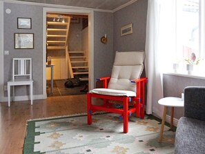 A room with a wooden floor, a patterned rug, a white chair, a red and blue chair, a small table, and a window with a view. - 6 Person Holiday Home in Atvidaberg (Atvidaberg)