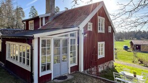 Punainen puutalo, jossa on suuri ikkunallinen kuisti, savupiippu ja pieni puutarha-alue.