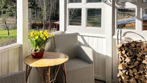 Un porche abrité avec un fauteuil en osier, une petite table ronde ornée d’un pot de jonquilles et une pile de bois de chauffage.