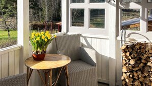 Un porche abrité avec un fauteuil en osier, une petite table ronde ornée d’un pot de jonquilles et une pile de bois de chauffage.