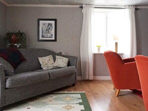 Ein Wohnzimmer mit einem grauen Sofa, einem roten Sessel, einem gemusterten Teppich, einem gerahmten Bild an der Wand und einem Fenster mit Vorhängen.