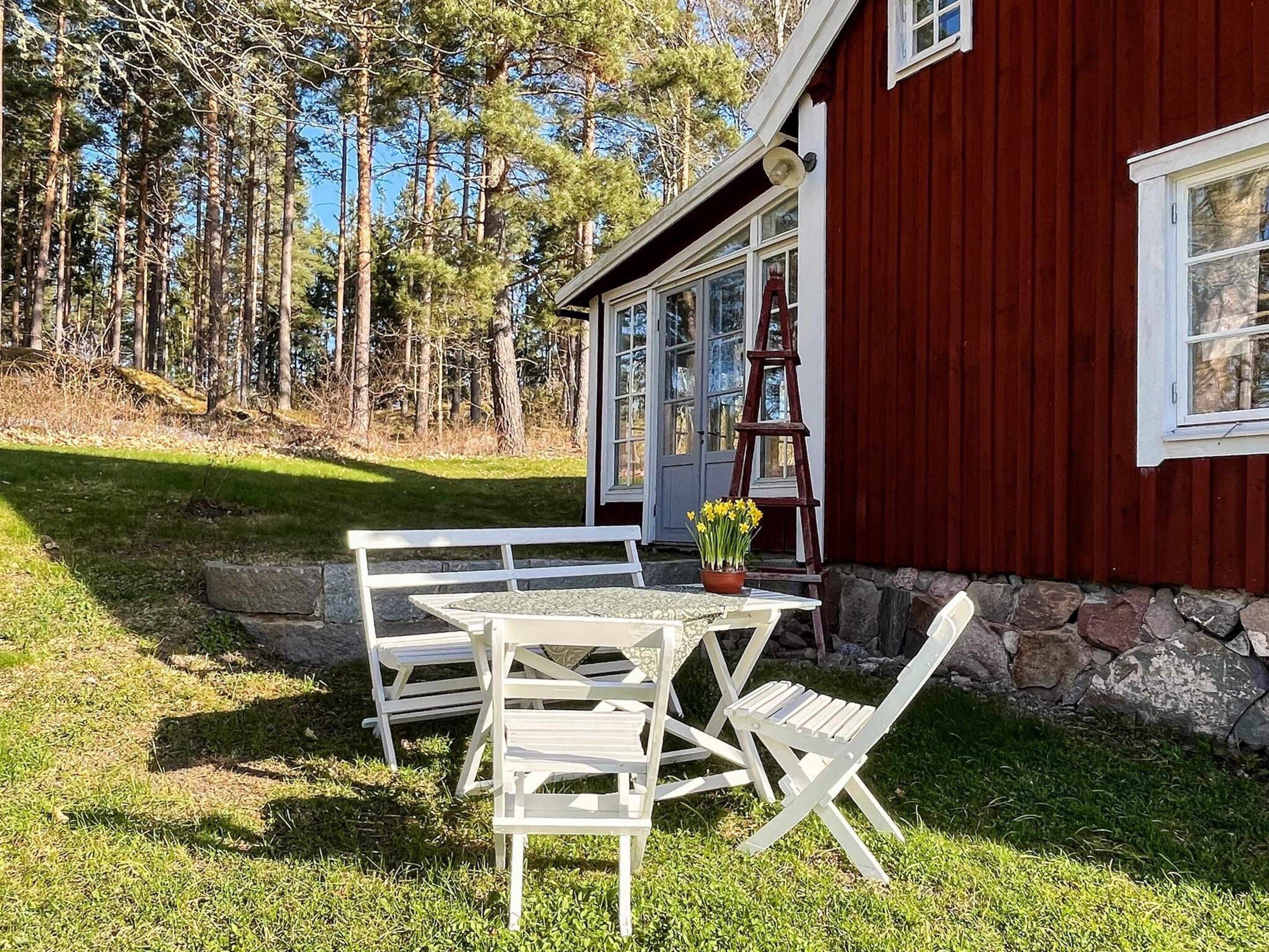 A red building with a stone foundation and a white door. There is a white table and two white chairs outside.