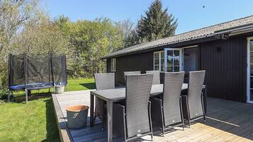 Eine Holzterrasse mit Gartenmöbeln, einem Trampolin und einem Gebäude mit dunkler Fassade und großen Fenstern.