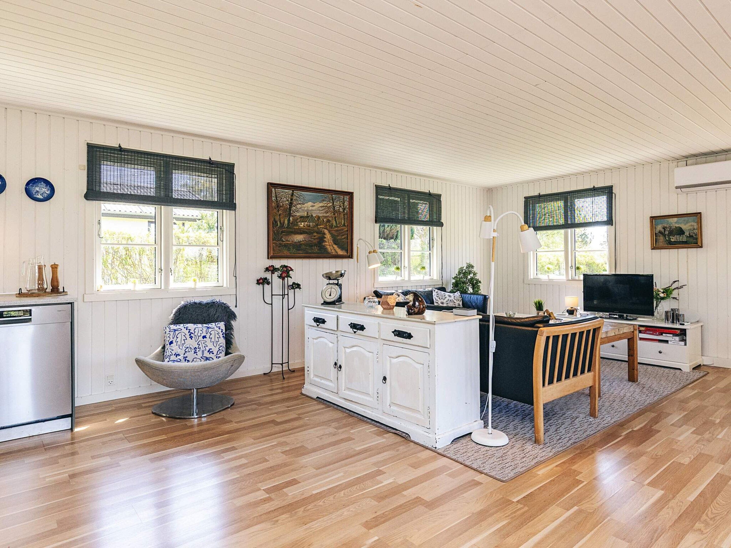 A room with a wooden floor, white cabinetry, a dining table with chairs, a sofa, a TV, and a window with a view of greenery.