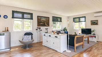 A room with a wooden floor, white cabinetry, a dining table with chairs, a sofa, a TV, and a window with a view of greenery.