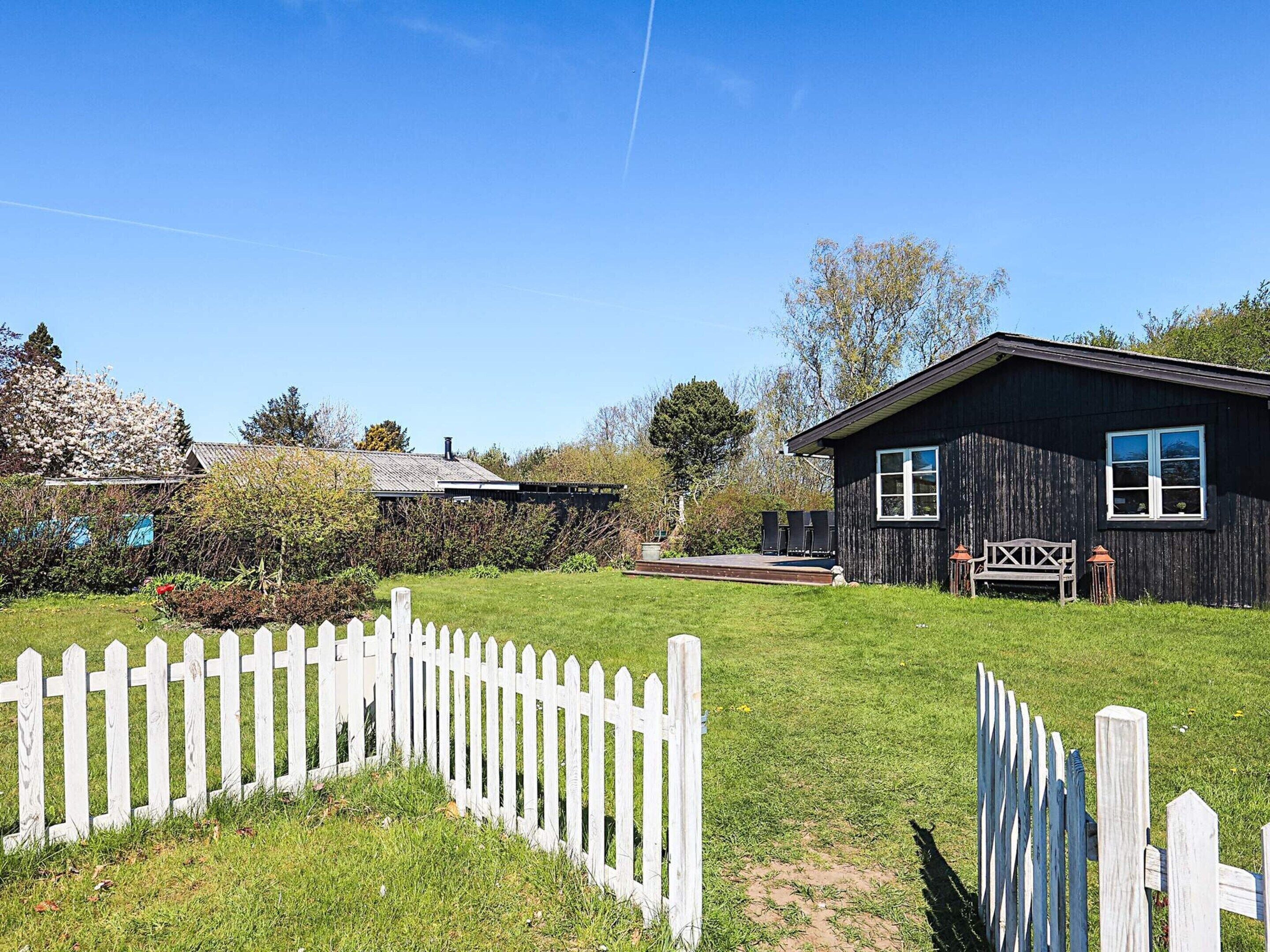 A wooden cabin with a deck, surrounded by a white picket fence, and a clear blue sky.