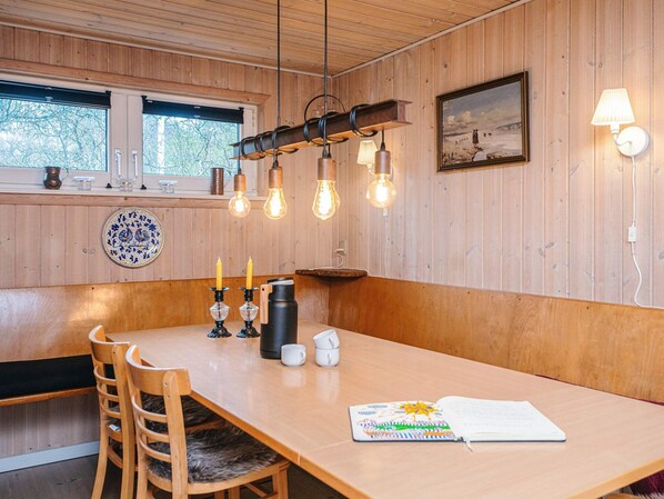 Un interior de madera con una mesa de comedor, sillas, lámparas colgantes, un candelabro, una cafetera y un cuadro en la pared.