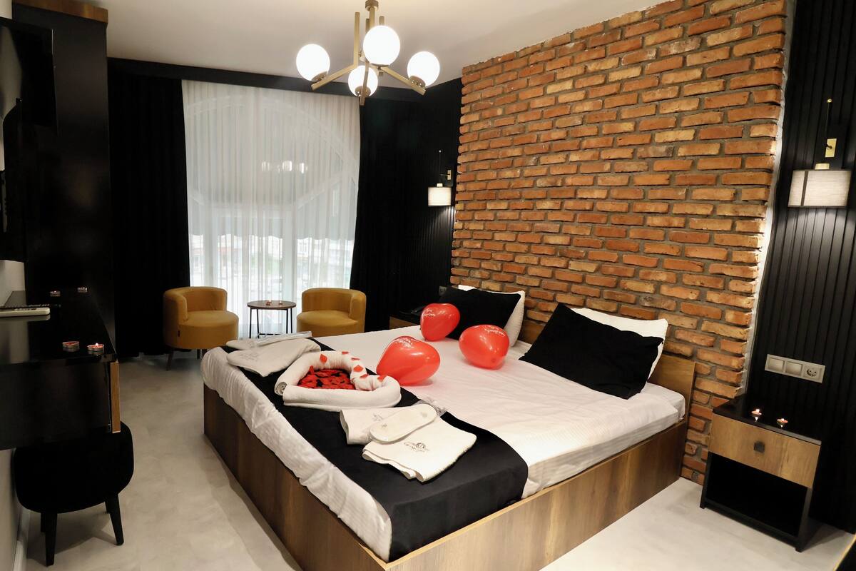 A modern hotel room with a brick accent wall, a large bed with white bedding, two armchairs, a small table, and a TV on a black stand.
