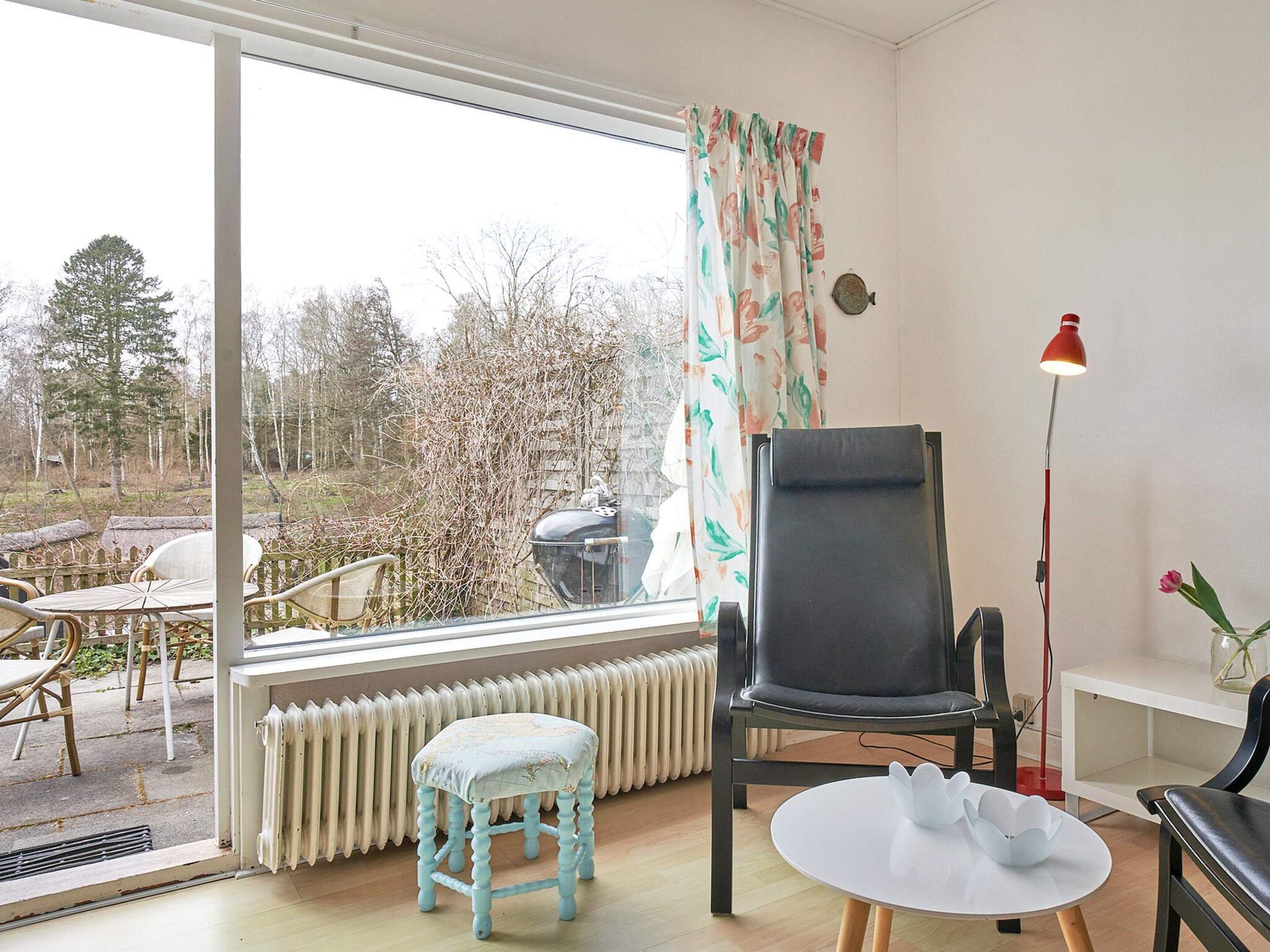 Une pièce avec une grande fenêtre, un radiateur, une chaise noire, un petit tabouret, une table blanche et un lampadaire rouge.