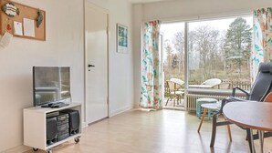 A room with a television on a cart, a door, a window with curtains, a table with chairs, and a view of the outdoors.