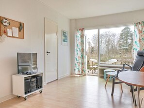 A room with a TV on a cart, a dining table with chairs, a window with curtains, and a door. - 2 Person Holiday Home in Aakirkeby (Akirkeby)