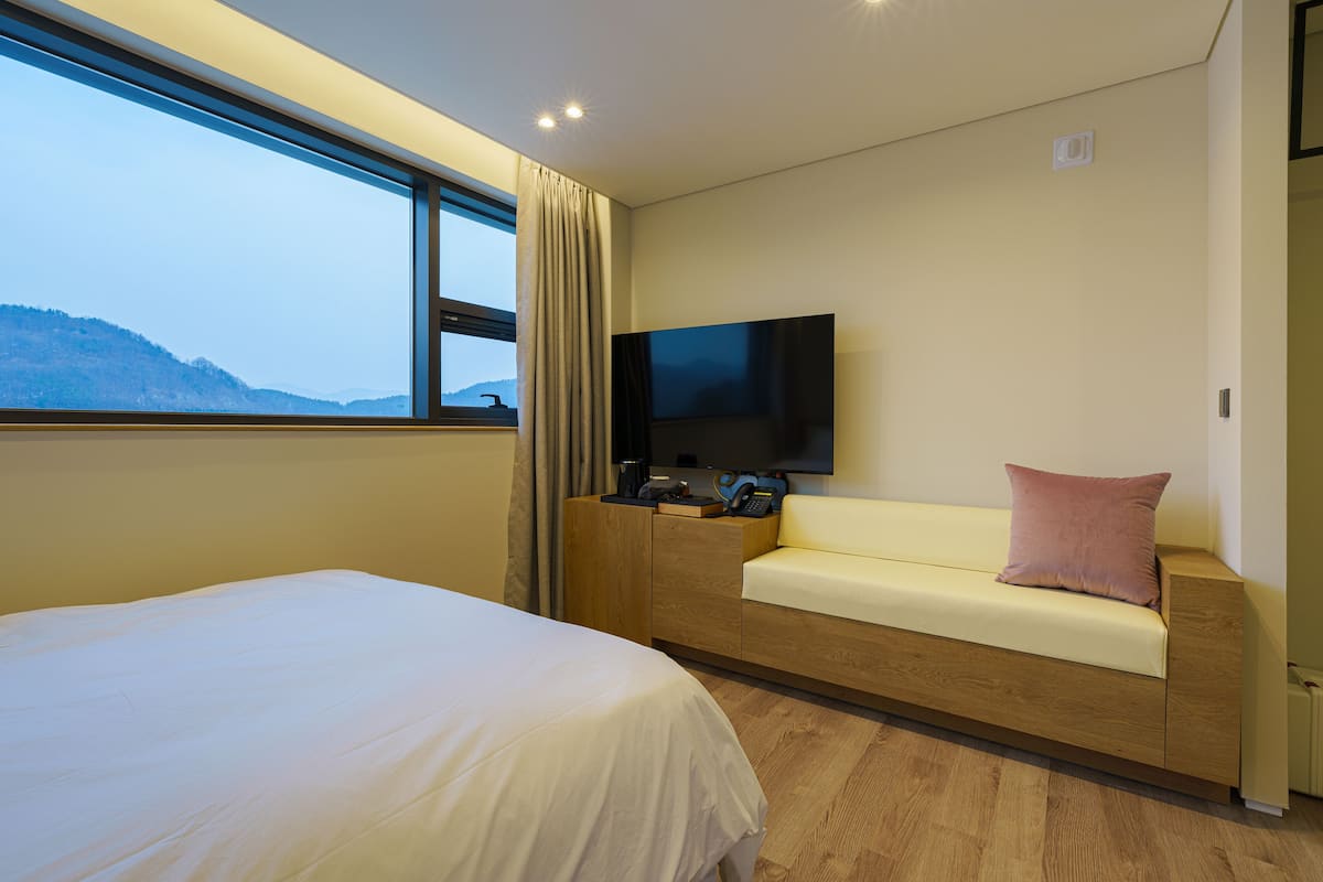 Une chambre d’hôtel moderne avec un grand lit, un canapé, un téléviseur fixé au mur et une vue sur les montagnes par la fenêtre.