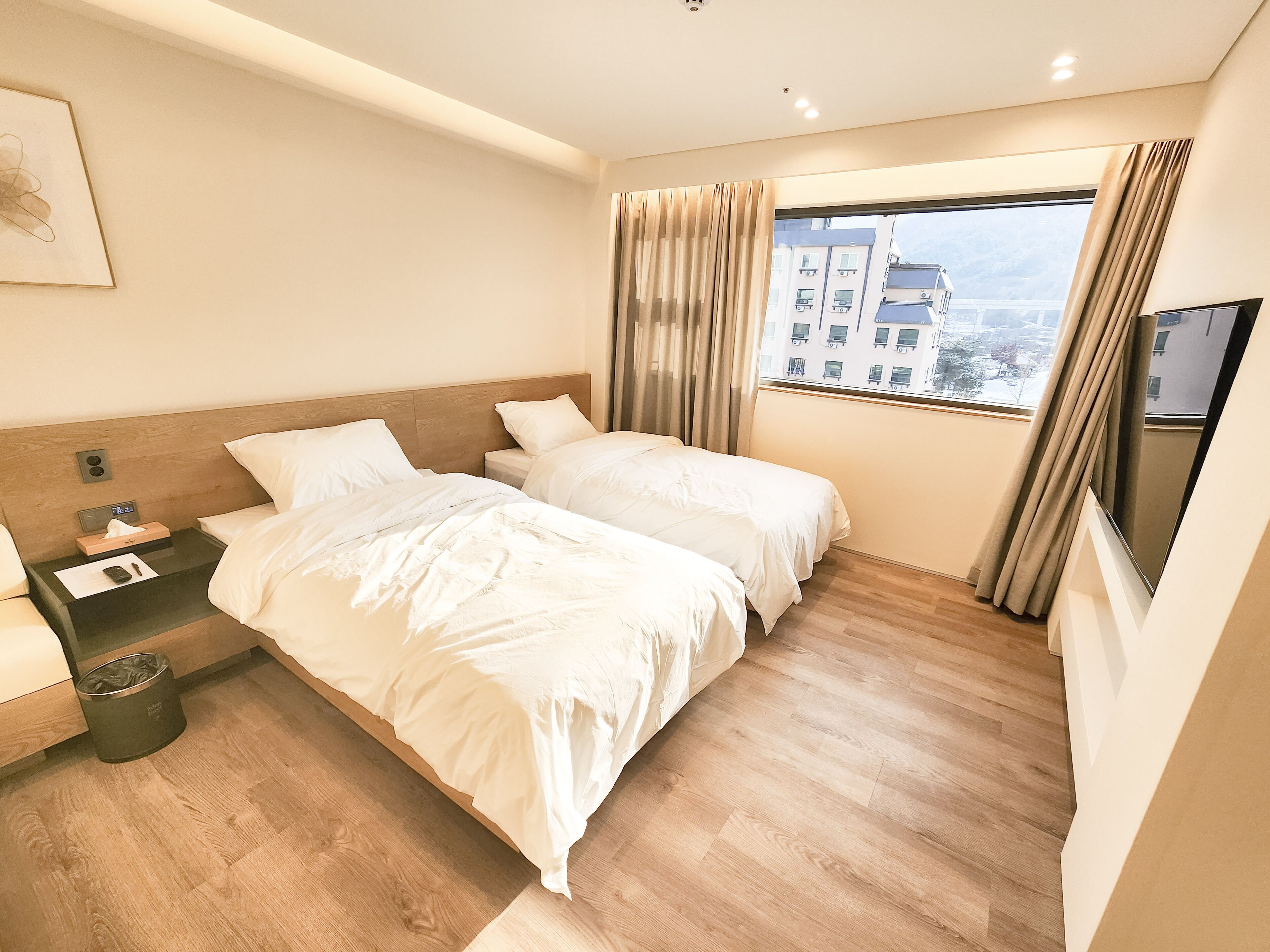 A hotel room with two beds, a bedside table, a TV, and a window with curtains.