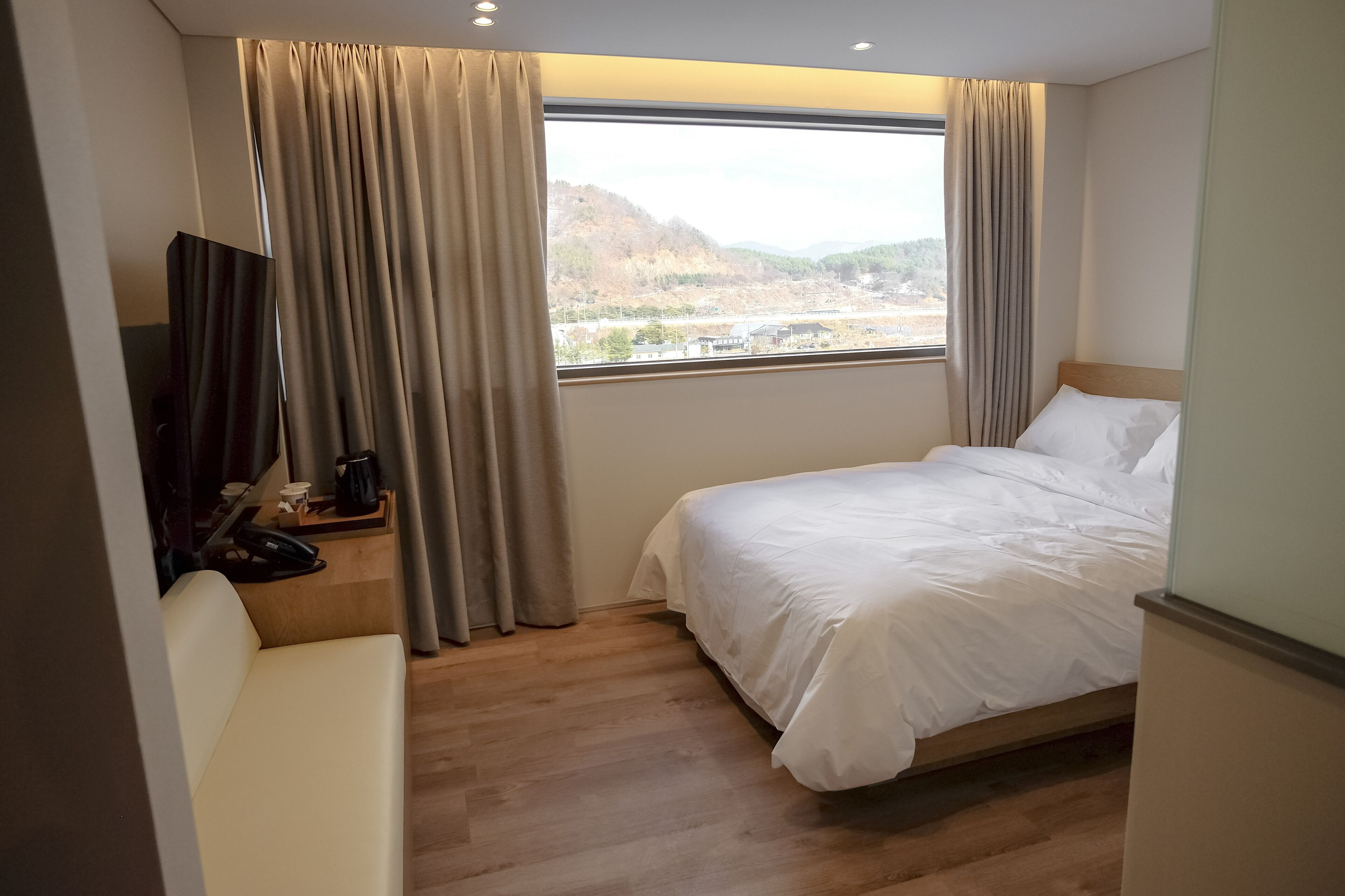 A hotel room with a bed, a desk with a phone and a coffee maker, and a view of a hilly landscape through a window with curtains.