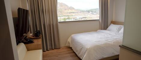 A hotel room with a bed, a desk with a phone and a coffee maker, and a view of a hilly landscape through a window with curtains.