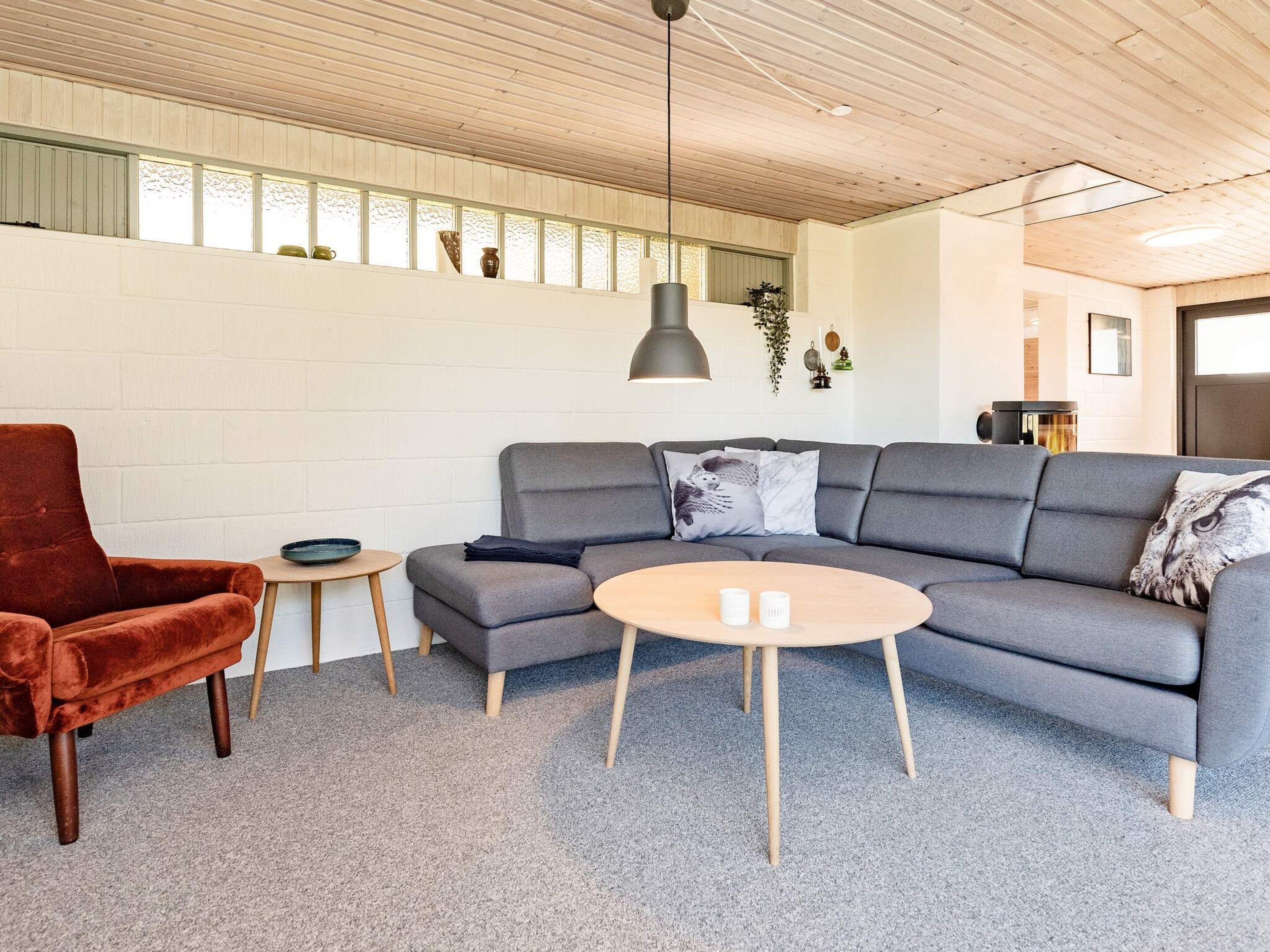 Uma sala de estar moderna com um sofá cinza, uma poltrona vermelha, uma mesa de centro redonda e teto de madeira.