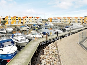 Uma marina com barcos atracados, uma passarela de madeira e uma fileira de casas coloridas ao fundo.