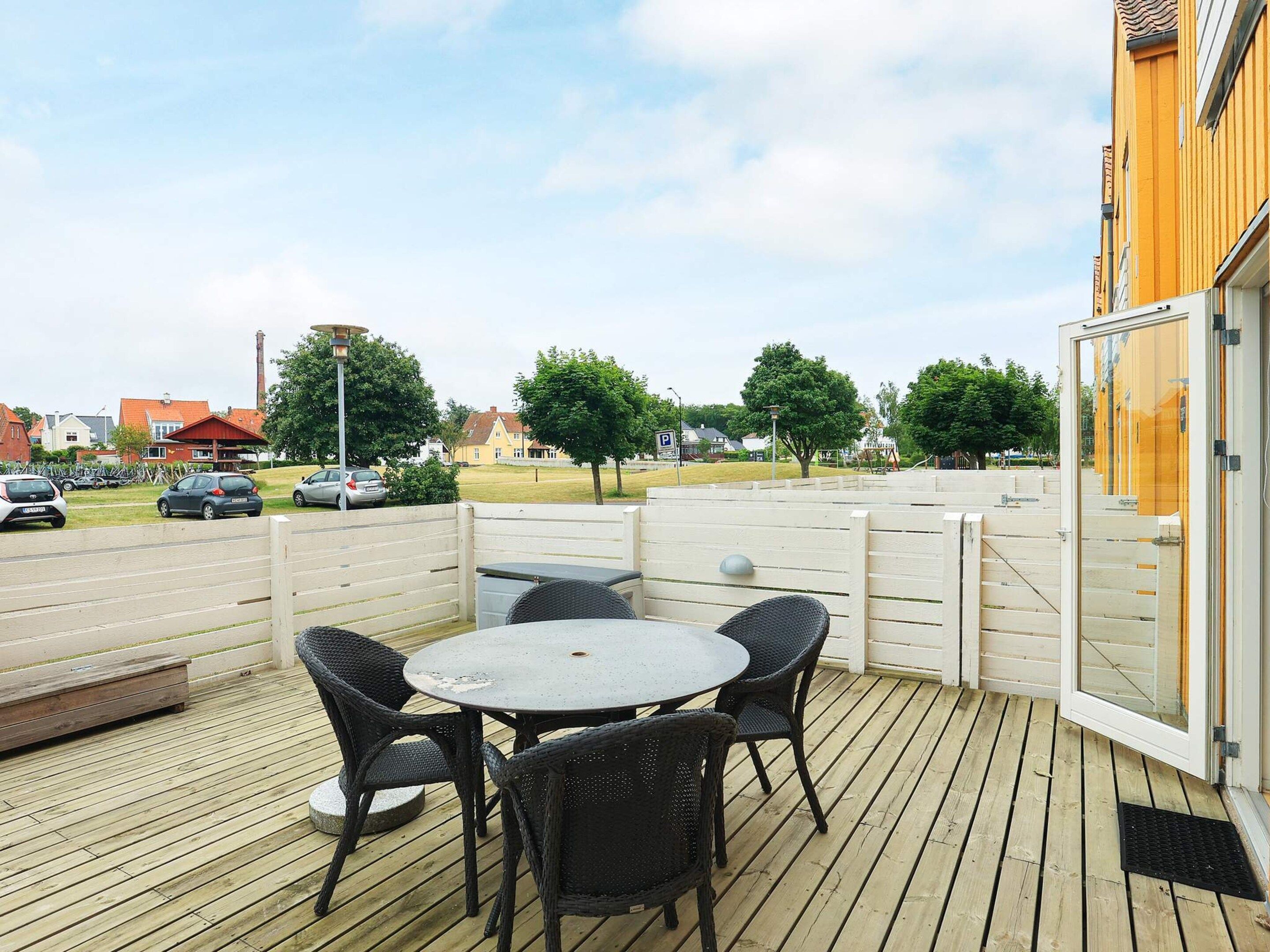 Un patio al aire libre con una mesa redonda y cuatro sillas, rodeado por una terraza de madera y una valla blanca.