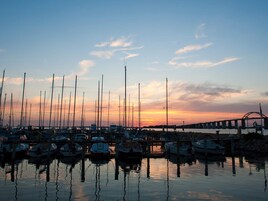 Un puerto deportivo con varias embarcaciones amarradas, un puente al fondo y un atardecer.