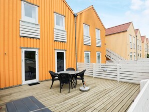 A wooden deck with outdoor furniture, a white fence, and a yellow building with multiple windows and doors.