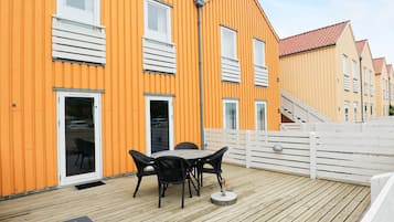 Eine Holzterrasse mit Gartenmöbeln, einem weißen Zaun und einem gelben Gebäude mit mehreren Fenstern und Türen.