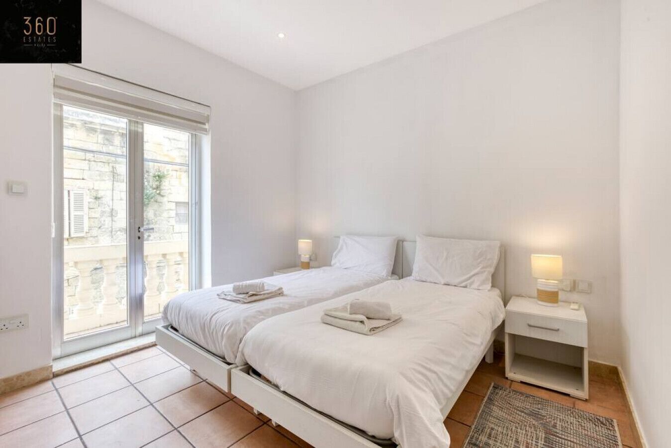 A bedroom with two beds, a window, and a nightstand with a lamp.