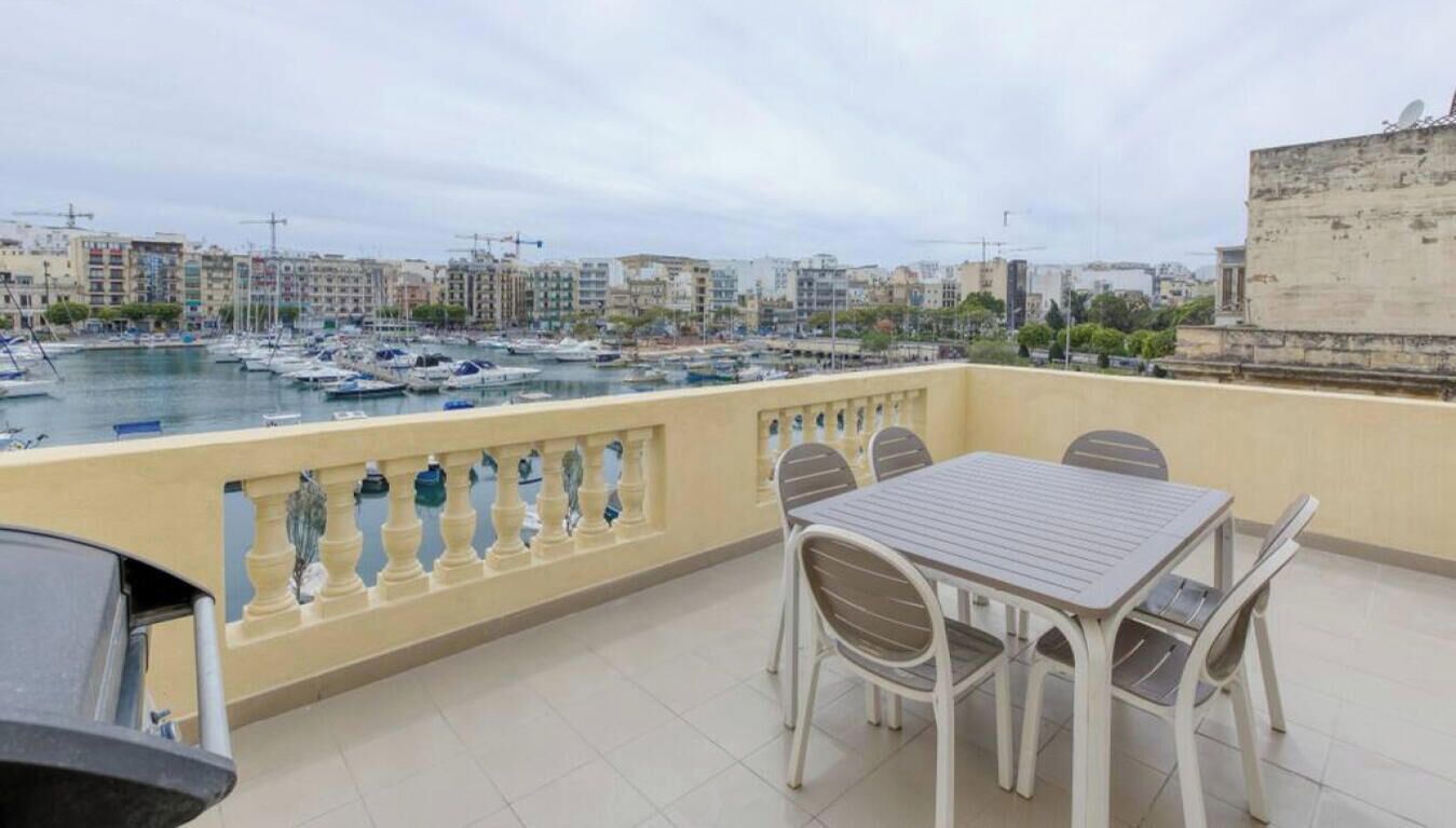A balcony with a table and chairs overlooking a marina with boats and buildings in the background.