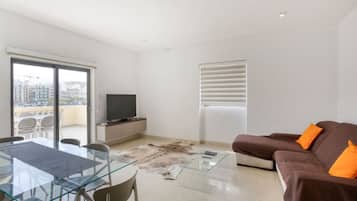 A modern living room with a glass dining table, brown sofa, and a television on a cabinet.