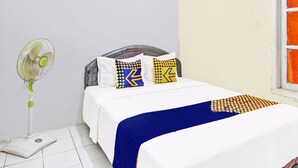 Una cama bien hecha con colcha blanca y azul, dos almohadas con diseños geométricos, un ventilador de pie y una balanza en el suelo.