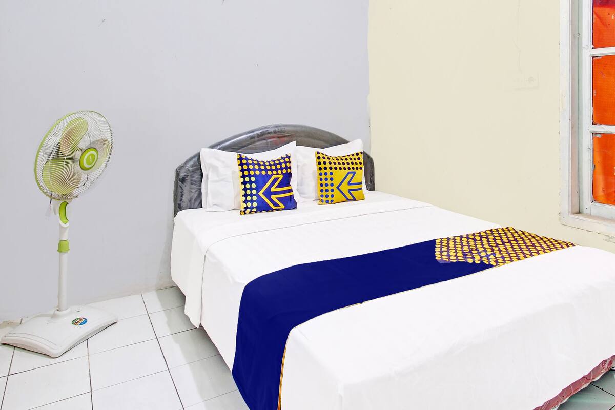 A neatly made bed with a white and blue bedspread, two pillows with geometric patterns, a standing fan, and a scale on the floor.