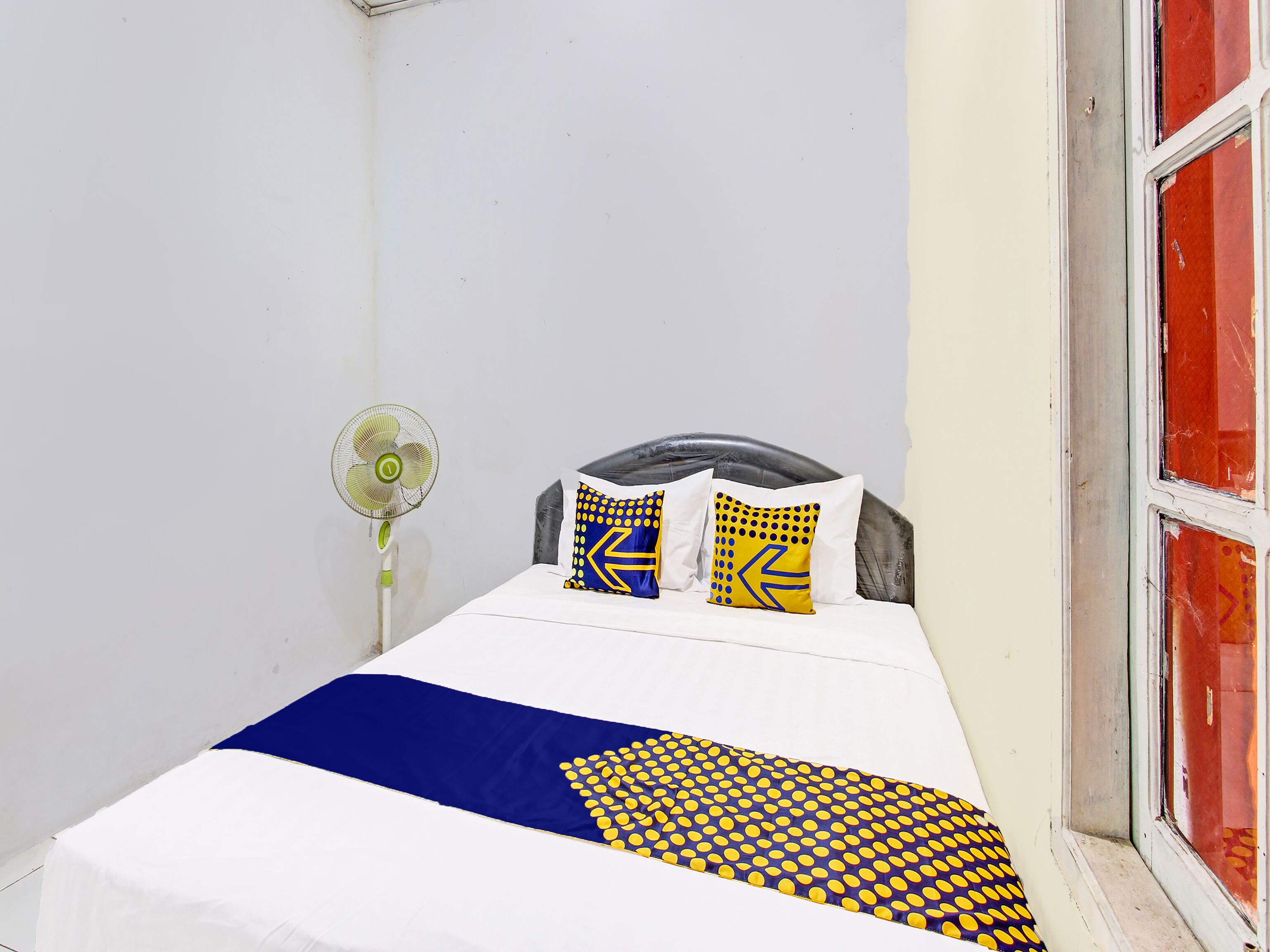 A bed with a white and blue bedspread, two pillows with yellow and blue patterns, and a standing fan.