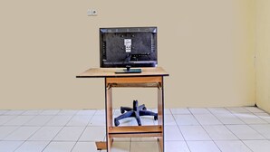 Un televisor sobre una mesa de madera en una habitación con suelo de baldosas blancas y paredes lisas.