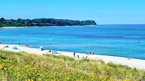 A beach with people swimming and relaxing, a clear sky, and a forested island in the background. - 6 Person Holiday Home in Gilleleje (Gilleleje)